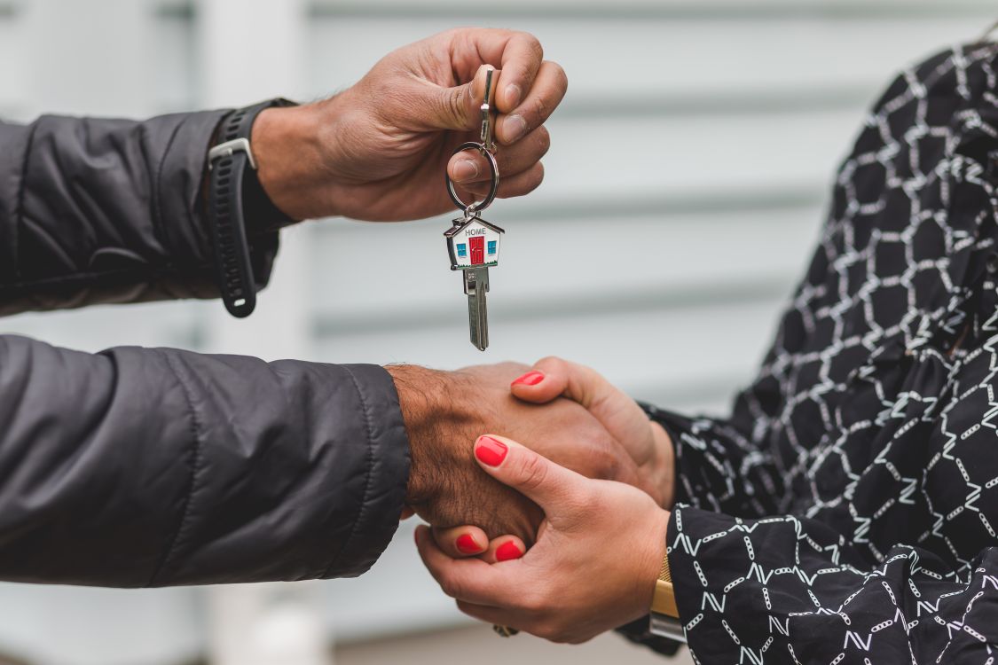 Close-up of a real estate agent's hand giving a set of house keys to a new homeowner in front of a welcoming suburban house.