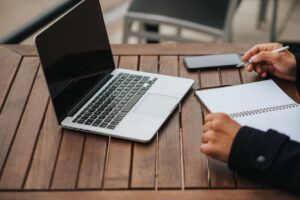 Professionally dressed person sits at a modern, bright desk, looking thoughtfully between a laptop and a physical notebook...