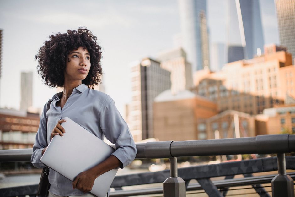 A well-dressed professional looks out a large office window at a bright city skyline, contemplating their future career in real estate.